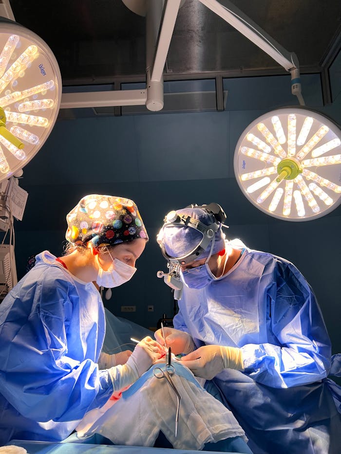 about-us Two surgeons in blue scrubs performing surgery under bright, focused lights in an operating room.